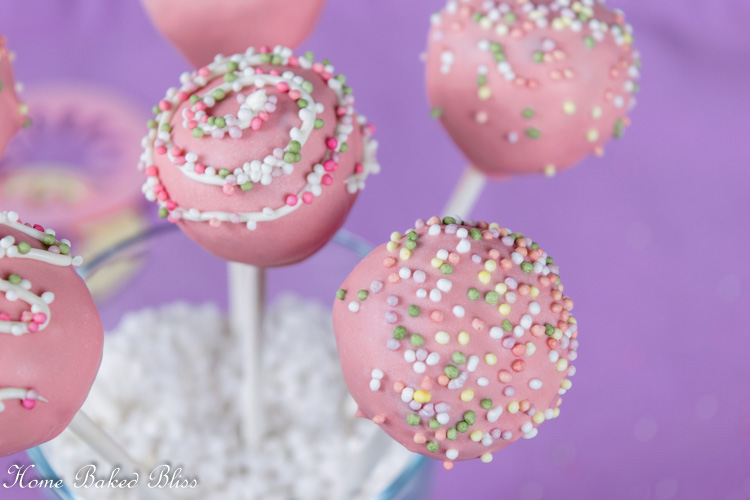 Chocolate cake pops with pink coating and colorful sprinkles in a vase filled with white stones.