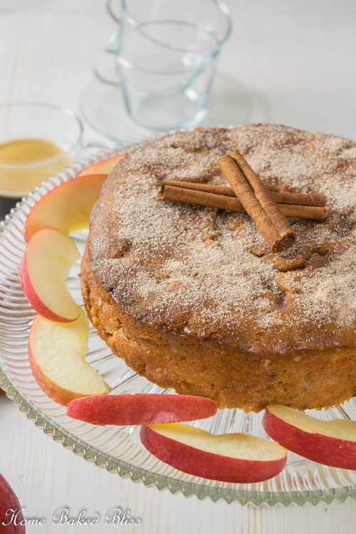 Cinnamon apple cake on a glass cake stand next to a cup of coffee and apple slices.