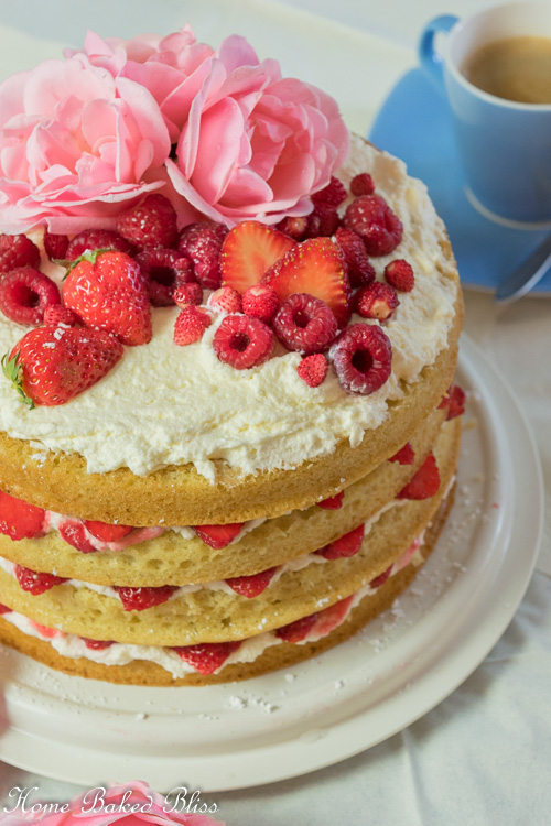 Berry Layer Cake garnished with berries and roses beside a cup of coffee.