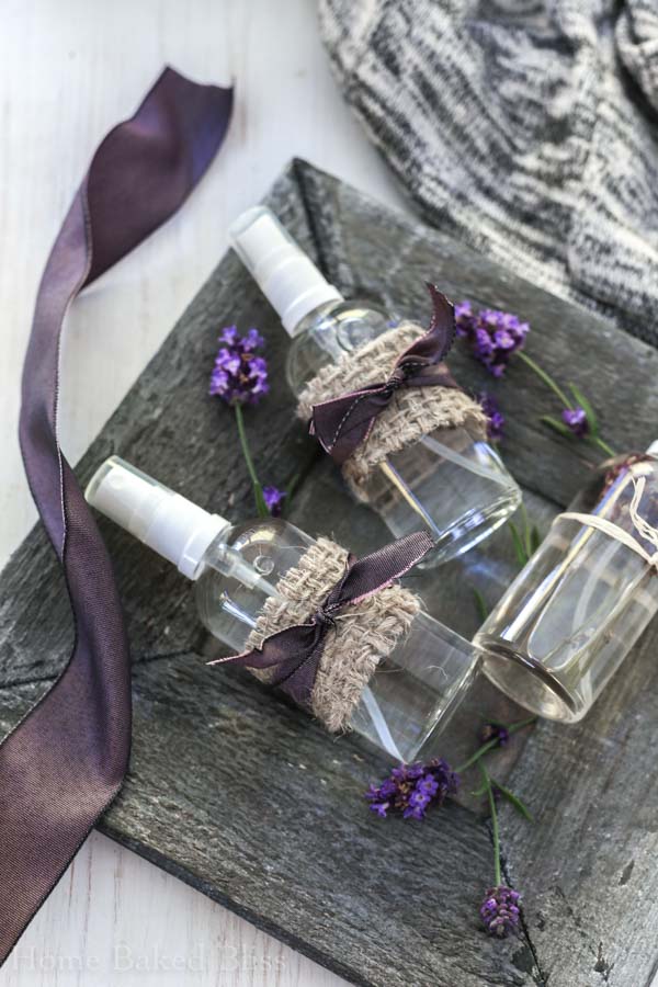 Three bottles of lavender linen spray on a wooden plate next to a purple ribbon and lavender buds.