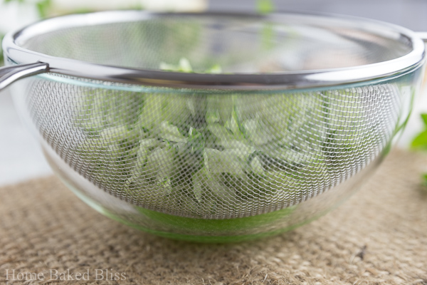 A glass bowl with a sieve on top with grated cucumber inside.