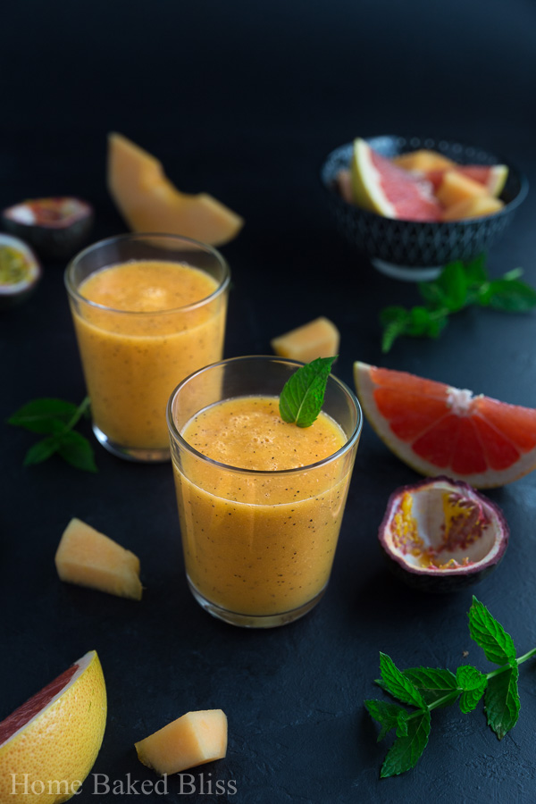 Two glasses of tropical smoothie garnished with a mint leaf beside various fruits