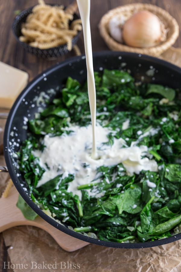 Heavy cream being poured into a skillet filled with roasted spinach