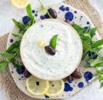 Greek tzatziki in a bowl on a blue plate garnished with olives and mint.