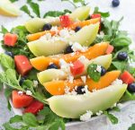 A melon salad on a bed of arugula, topped off with feta cheese and blueberries on a white plate.