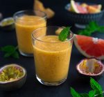 Two glasses of tropical smoothie garnished with a mint leaf beside various fruits
